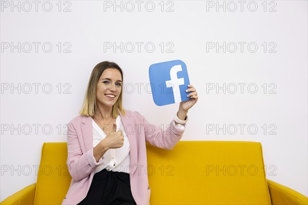 Young woman sitting yellow sofa holding facebook icon showing thumbup sign