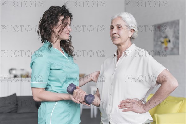 Beautiful old lady is doing exercise with dumbbell with help physiotherapist