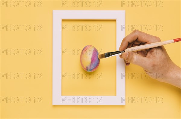 Close up hand painting easter egg with brush inside frame yellow background