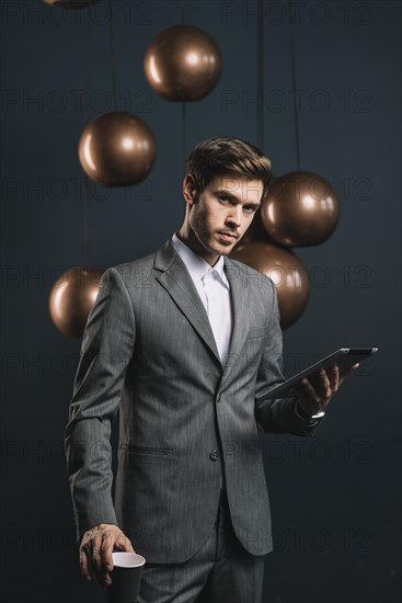 Confident young man holding disposable cup digital tablet against dark background