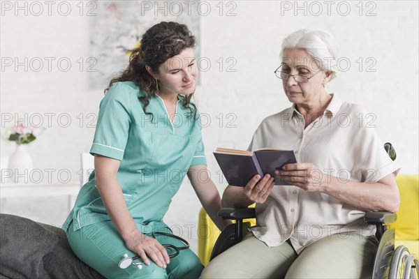 Disabled senior female patient sitting wheel chair reading book with nurse