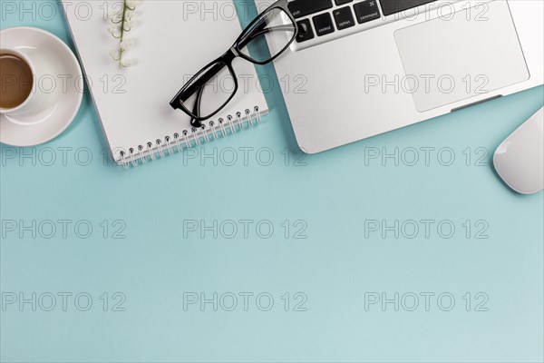 Elevated view coffee cup spiral notepad eyeglasses laptop mouse blue backdrop