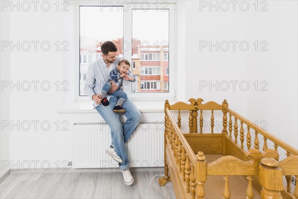 Father carrying his baby boy son sitting window sill looking wooden empty crib