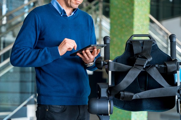 Unrecognizable computer engineer in mechanical exoskeleton looking at the robot with a tablet. Physiotherapy in a modern hospital: Robotic skeleton. Physiotherapy Rehabilitation Scientists