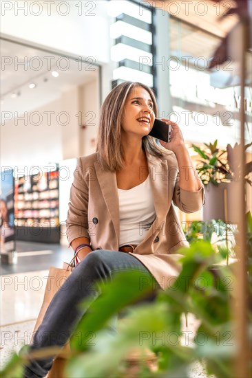 Happy woman talking to the phone sitting on a mall holding shopping bags and smiling looking away