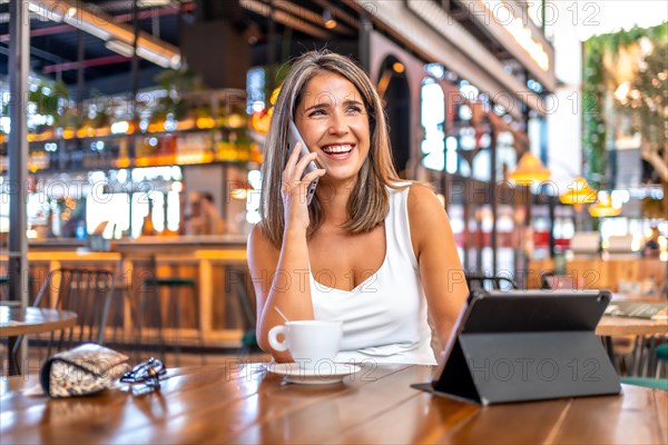 Distracted businesswoman talking to the phone in a cafeteria sitting with a digital tablet and a coffee cup