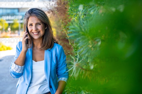 Smiling businesswoman talking to the phone next to a green area in the street