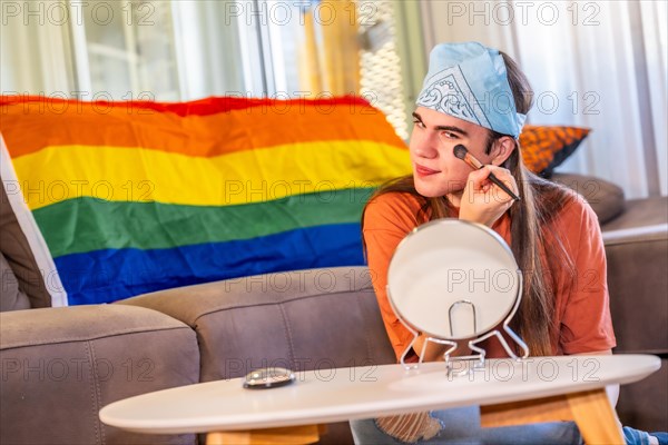 Transgender young man making up using brush and mirror sitting in the living room next to a rainbow lgbt flag at home