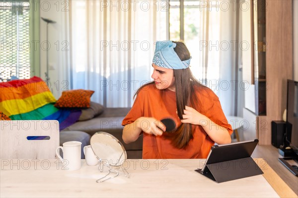 Distracted transgender person arranging the long hair at home sitting on a table with a digital tablet and a mirror