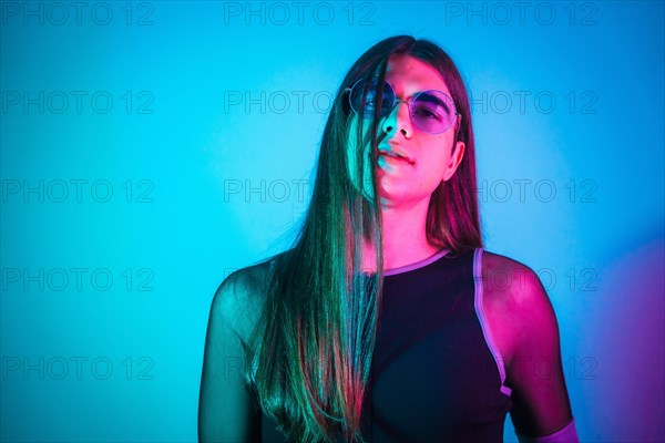 Portrait of a futuristic transgender person with long hair and sunglasses illuminated by neon lights