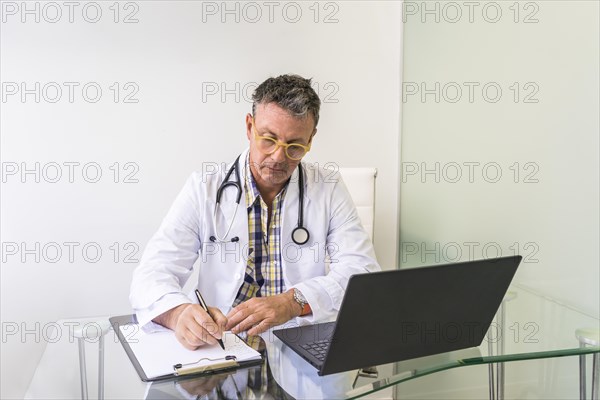 Male doctor working on laptop and clipboard in medical office