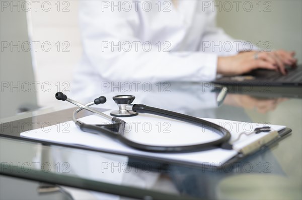 Close-up of a stethoscope and clipboard on desk while female doctor is writting on laptop