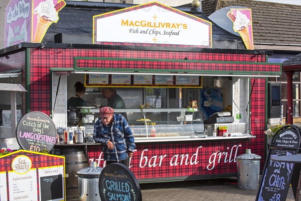 Old man in front of fish & chips and seafood stand in the city Oban