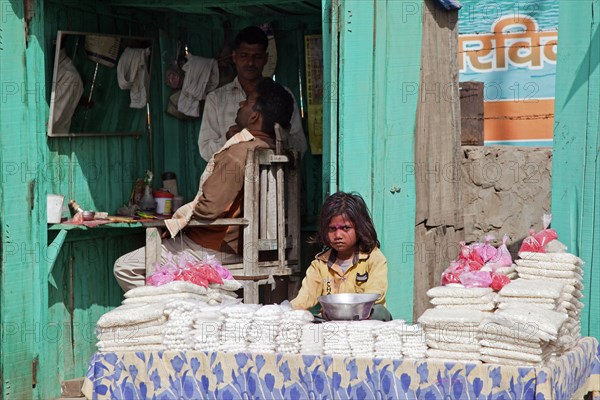 Hairdresser cutting hair in barbershop and child behind display selling goods in Govardhan