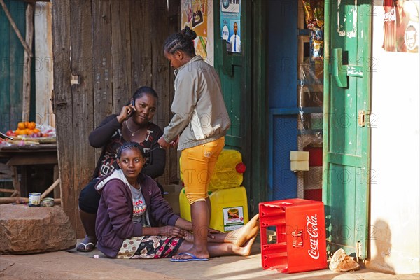 Malagasy woman on the phone and girls braiding each other's hair in front of shop in the city Ambalavao