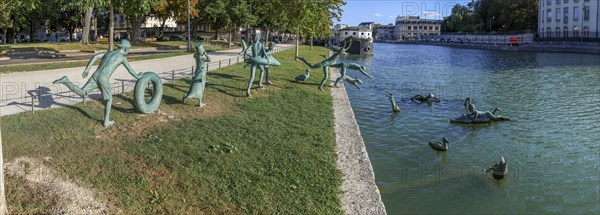 Panoramic photo of a group of bronze sculptures of playful children La Ribambelle Joyeuse by Belgian artist Tom Franzten on the banks and in the Canal du Trevois