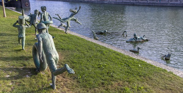 Panoramic photo of a group of bronze sculptures of playful children La Ribambelle Joyeuse by Belgian artist Tom Franzten on the banks and in the Canal du Trevois