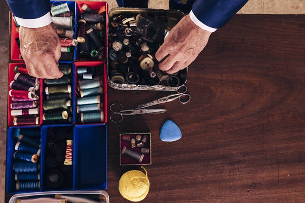 Overhead view male fashion designer s hand holding thread spool buttons from box wooden desk