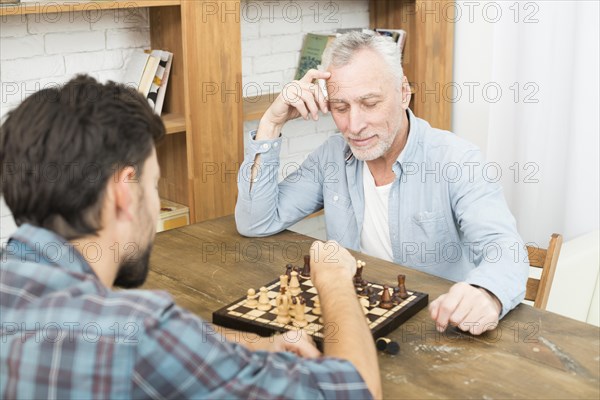 Pensive aged man young guy playing chess table near bookshelves