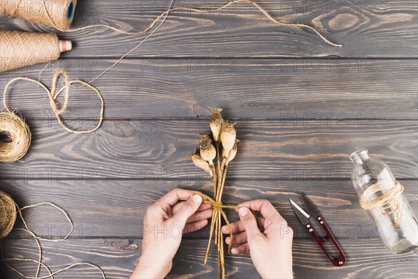 Person s hand tying dry poppy pods with string near cutter glass bottle desk