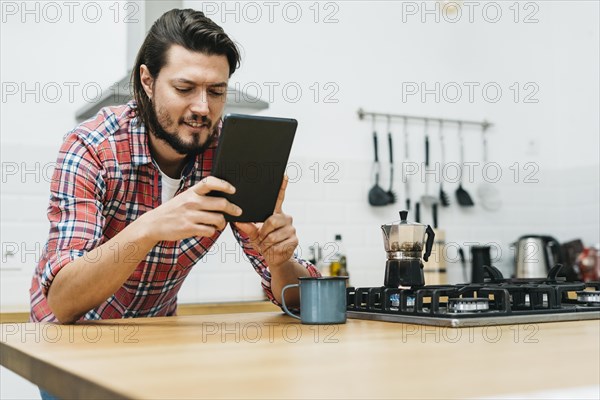 Portrait smiling young man leaning wooden kitchen counter looking smart phone
