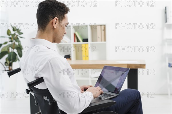 Side view disabled young man sitting wheelchair using laptop office
