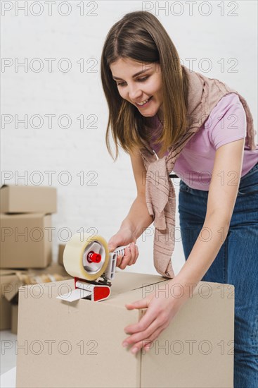 Smiling young woman holding packing machine sealing cardboard boxes with duct tape