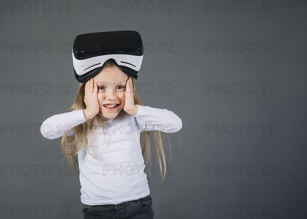 Surprised girl wearing virtual reality glasses her head touching her cheeks against gray background