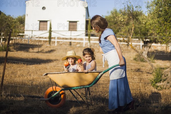 Two sister holding red apple sitting wheelbarrow being pushed by woman