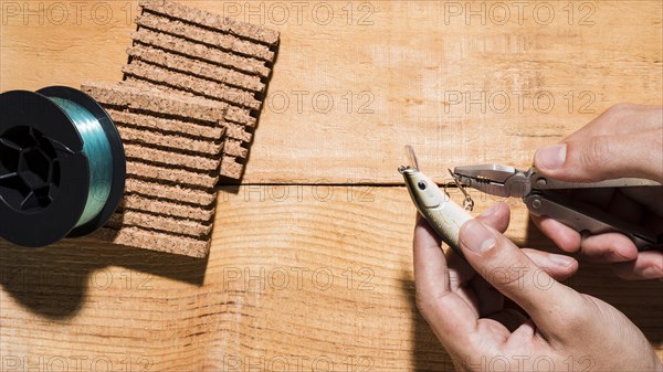 Close up man fixing hook with plier near reel cork board wooden desk