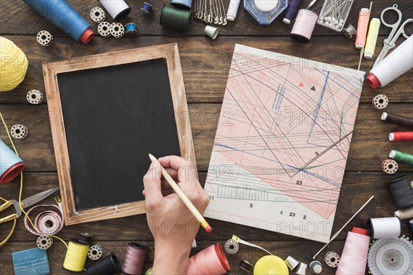 Crop hand drawing blackboard near sewing stuff