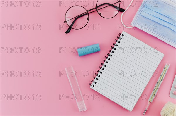 Eyeglasses thermometer surgical mask spiral notebook pink background