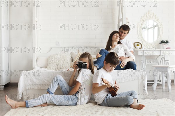 Loving parent sitting bed their daughter holding camera son playing ukulele