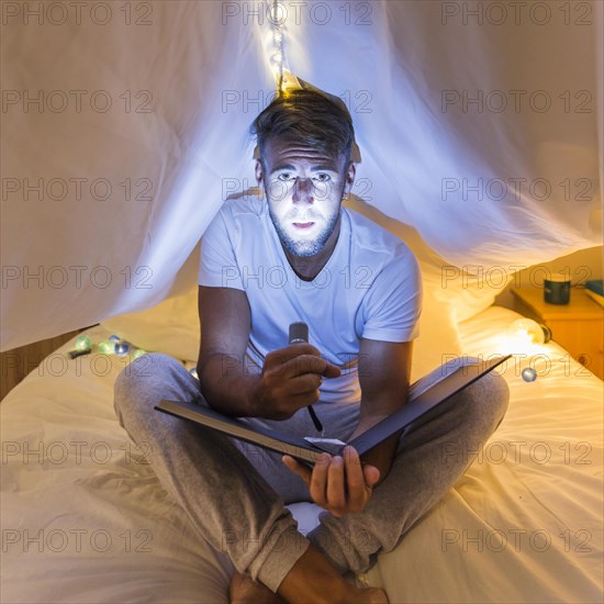 Man sitting curtain bed holding album highlights his face with flashlight