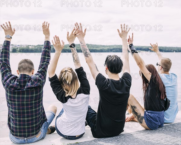 Rear view friends sitting near lake raising their hands