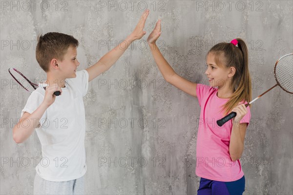Side view boy girl holding racket hand giving high five standing against wall