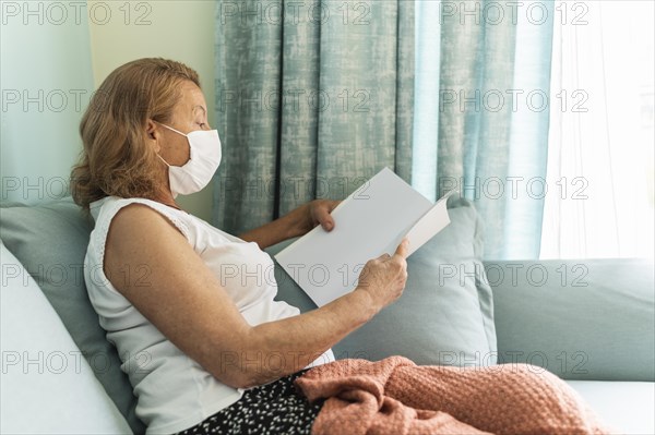Side view elder woman with medical mask home during pandemic reading book