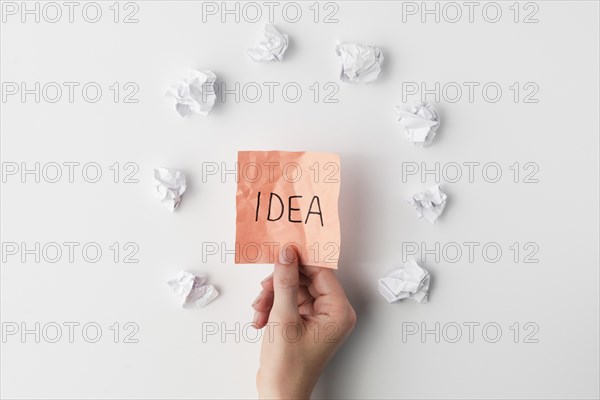 Top view human hand holding sticky note with idea text surrounded by crumpled paper white background