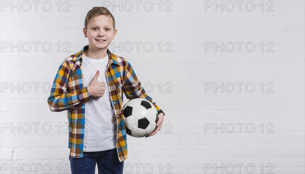 Smiling boy holding soccer hand showing thumb up sign standing against white brick wall