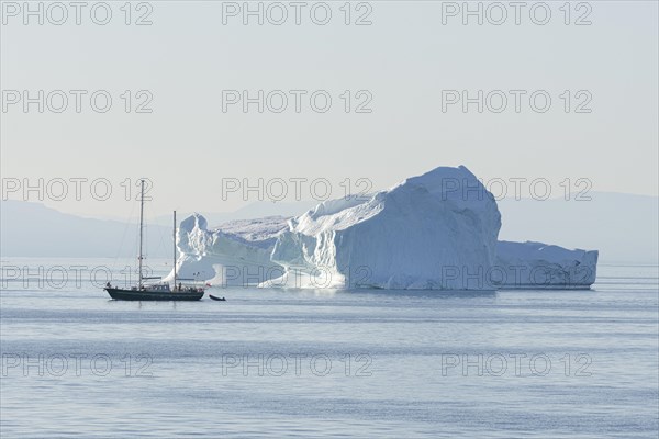 A small sailing ship passes an iceberg in the evening sun. Disko Bay