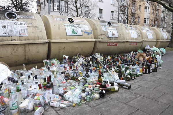 Overflowing recycling glass bottle containers with rows of bottles lined up in front on the footpath