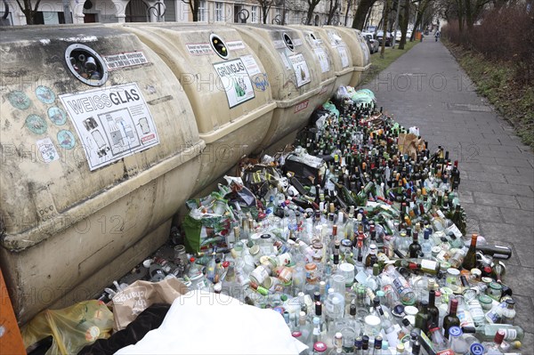 Overflowing recycling glass bottle containers with rows of bottles lined up in front on the footpath