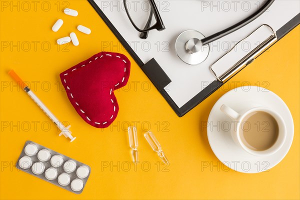 Red stitched heart coffee cup stethoscope eyeglass injection blister packed medicine clipboard against yellow backdrop