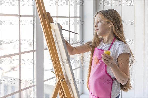 Side view girl holding yellow paint bottle hand painting easel with paintbrush