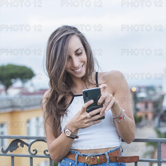 Smiling beautiful young woman looking mobile phone standing balcony