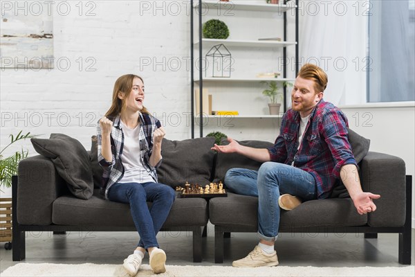 Young woman clenching her fist with joy looking his boyfriend shrugging while playing chess