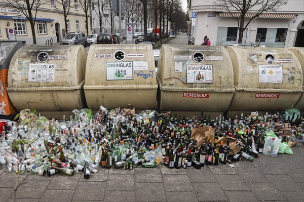 Overflowing recycling glass bottle containers with rows of bottles lined up in front on the footpath