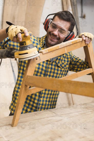 Smiling male carpenter softening edges wooden furniture with sander