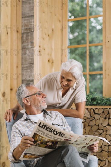 Smiling senior woman looking her husband sitting chair holding newspaper