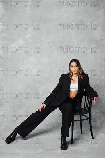 Stylish young woman in black pantsuit and ankle boots with rough sole sitting on chair put one leg aside